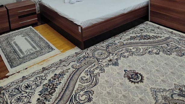 A bedroom with a bed, wooden wardrobe, a radiator, a small window, and a patterned rug. - DDS Hostel (Tashkent)