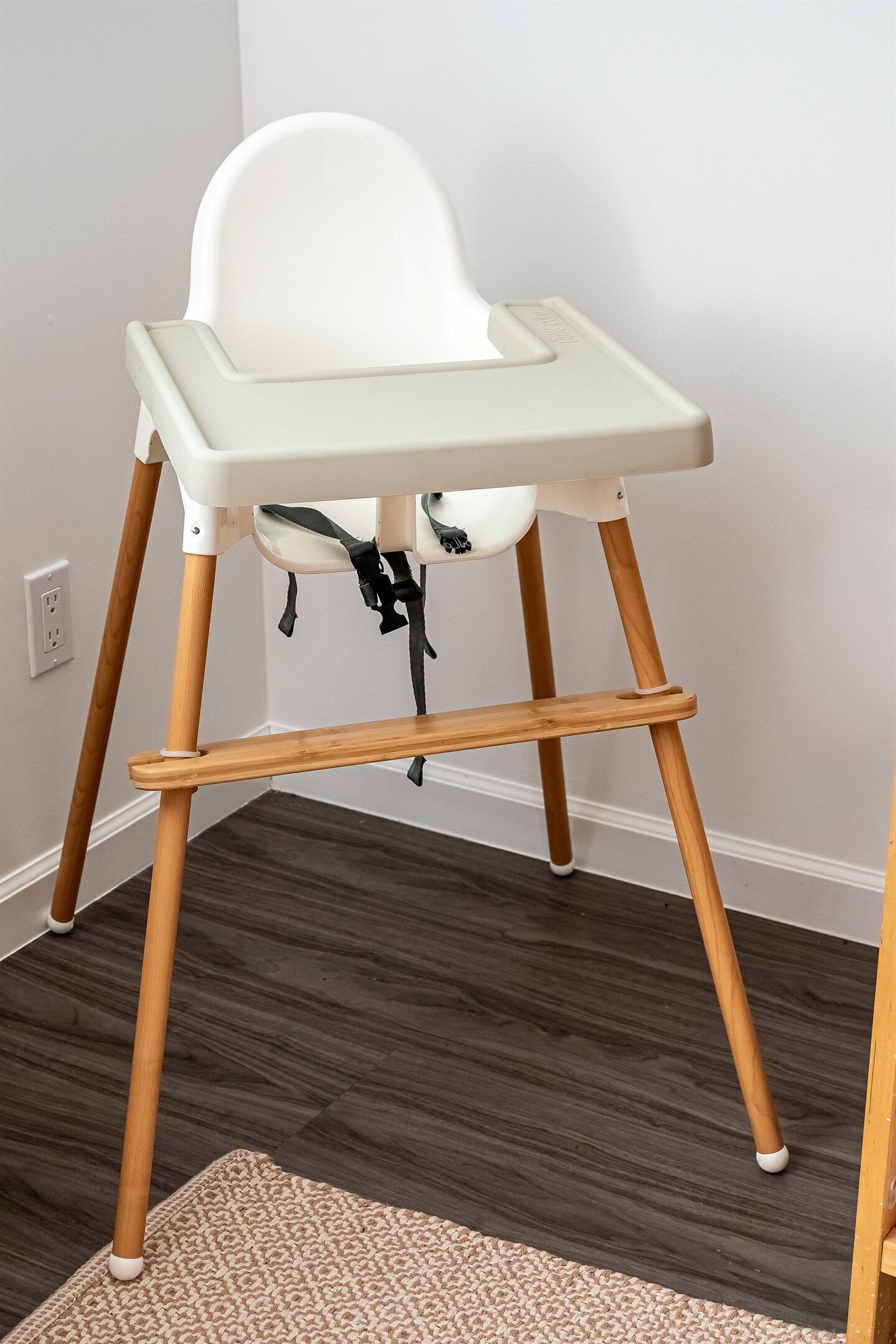 A high chair with wooden legs and a white seat, placed on a dark wooden floor with a beige rug in front.