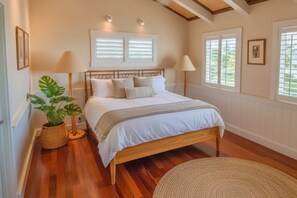 Een slaapkamer met een houten bed, twee bedlampen, een kamerplant en een groot raam met jaloezieën.