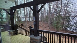A wooden deck with a stone fireplace, a black railing, and a view of leafless trees.