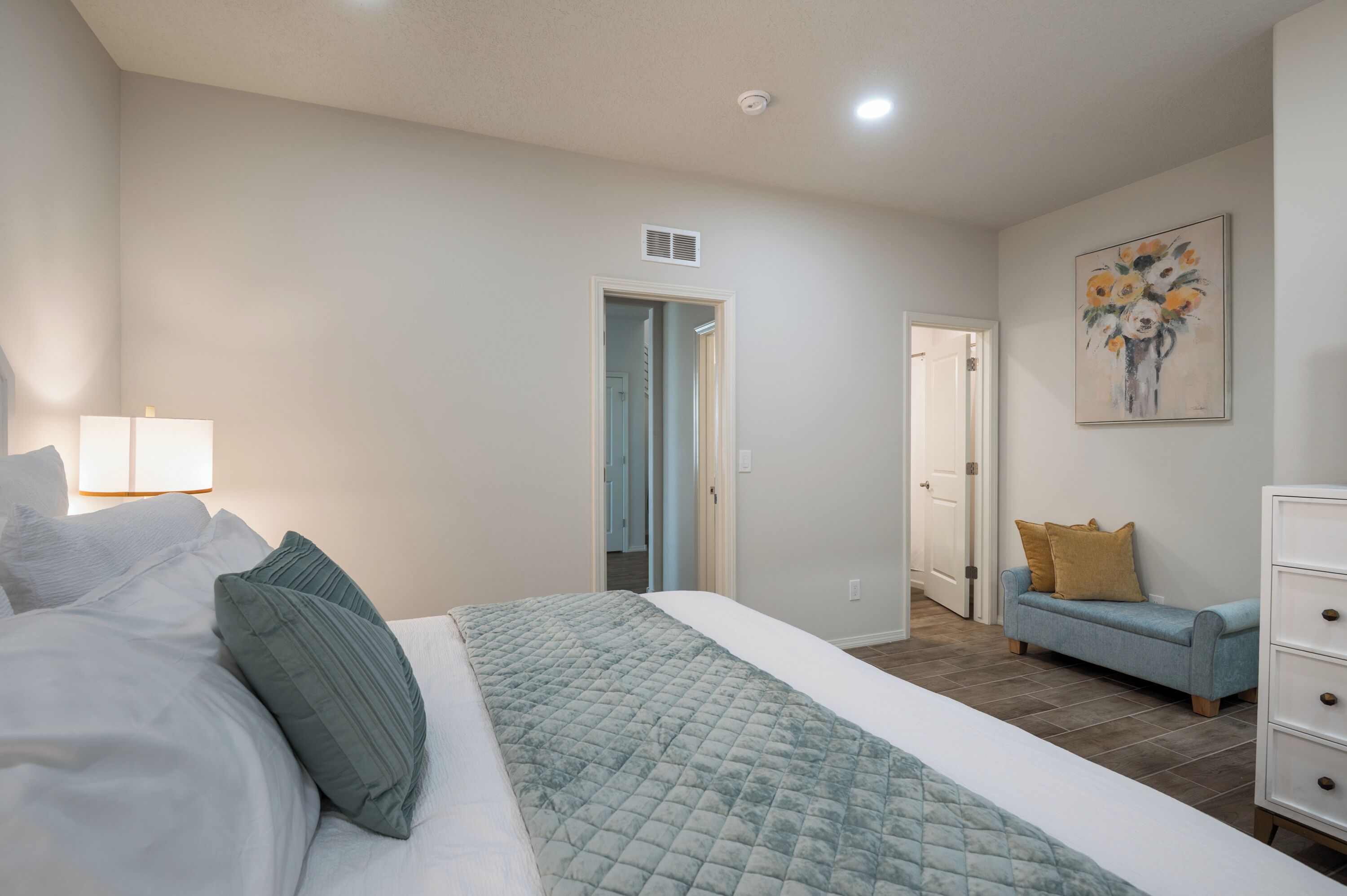 A bedroom with a bed, a nightstand, a dresser, a painting, and a door leading to another room.