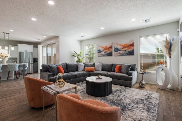 A modern living room with a sectional sofa, a coffee table, and orange armchairs. There is a dining area with a white table and chairs, and a kitchen in the background. - Spacious Family Friendly 5BR Retreat Sandia Views Movie Lounge + Fenced Yard (Albuquerque)