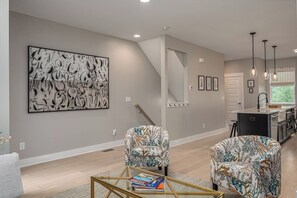 A modern living room with a large abstract painting, two patterned armchairs, a glass coffee table with books, and a kitchen area with pendant lighting.