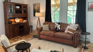 A living room with a brown leather sofa, wooden cabinet, and a large window with curtains. - Brand-new, PRIVATE 3-bed mountain retreat w/ hiking trails, HOT TUB, fire pit! (Pownal)