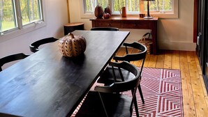 A dining area with a wooden table, chairs, and a rug. There are windows with a view of trees. - Brand-new, PRIVATE 3-bed mountain retreat w/ hiking trails, HOT TUB, fire pit! (Pownal)