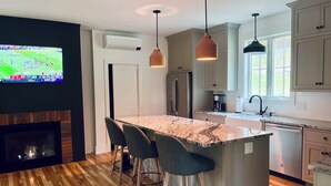 A modern kitchen with a marble island, bar stools, stainless steel appliances, and a flat-screen TV on the wall. - Brand-new, PRIVATE 3-bed mountain retreat w/ hiking trails, HOT TUB, fire pit! (Pownal)