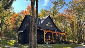 A cabin with a porch, surrounded by trees with autumn foliage. - Brand-new, PRIVATE 3-bed mountain retreat w/ hiking trails, HOT TUB, fire pit! (Pownal)