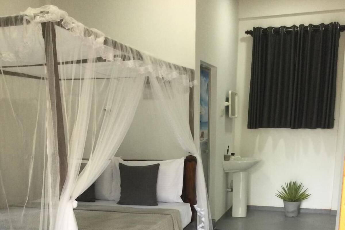 A bedroom with a four-poster bed, a bedside table, a small potted plant, and a window with dark curtains.