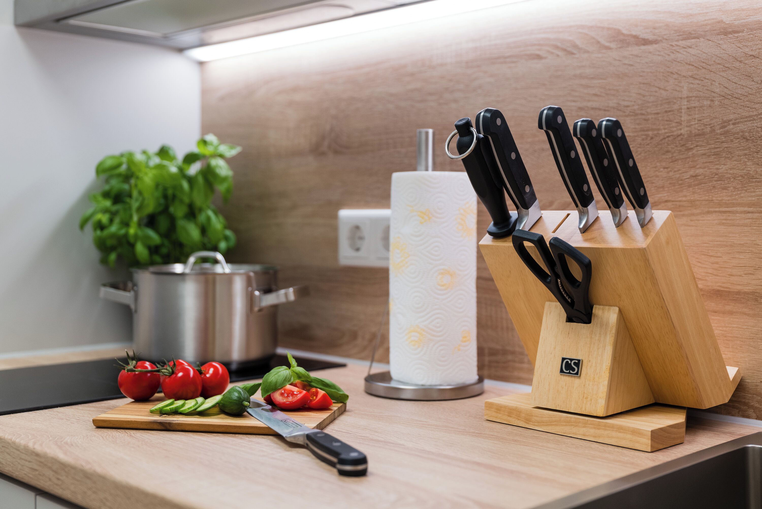 Un plan de travail de cuisine avec une planche à découper, des tomates, du basilic, un couteau, un porte-essuie-tout et une casserole.