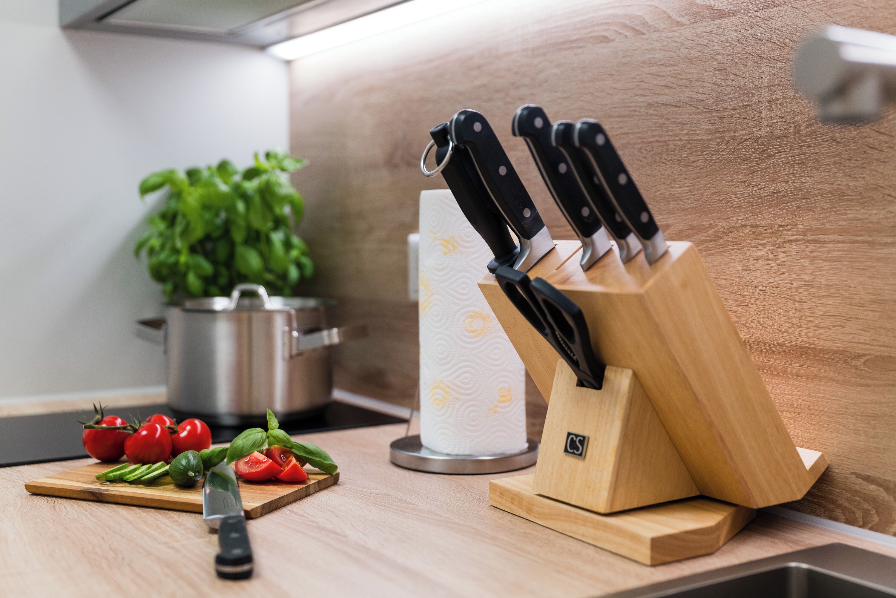 A kitchen counter with a cutting board, tomatoes, basil, and a knife block.
