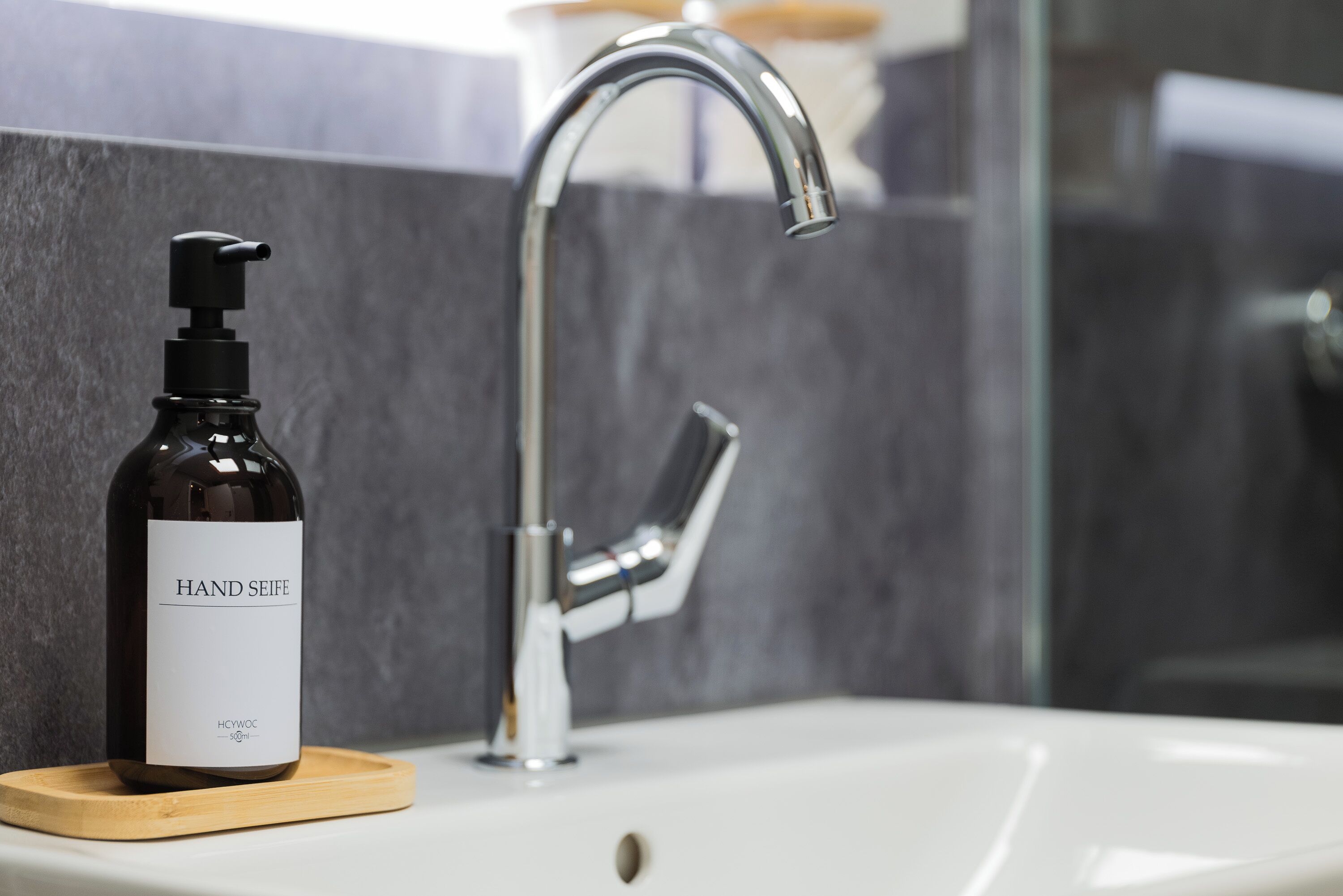 Ein Badezimmer mit einem weißen Waschbecken, einem modernen Wasserhahn und einer Handdesinfektionsmittel-Flasche auf einem Holzuntersetzer.