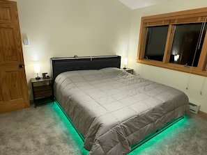 Een slaapkamer met een bed, nachtkastjes, een houten deur, een raam met jaloezieën en verlichting onder het bed in groen.