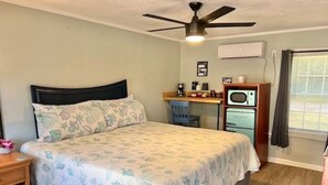 Une chambre à coucher comprenant un lit, un bureau, une chaise, un ventilateur de plafond, un four à micro-ondes et une fenêtre avec des rideaux.