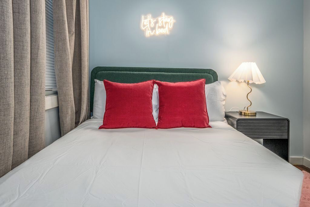 A neatly made bed with white bedding and two red pillows, a bedside lamp, and a neon sign on the wall.