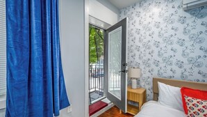 Une chambre avec un plancher en bois, un lit avec des oreillers blancs et rouges, une table de chevet avec une lampe, et une porte donnant sur un balcon.