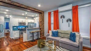 Un salon moderne avec un canapé gris, des rideaux orange et une cuisine en arrière-plan.