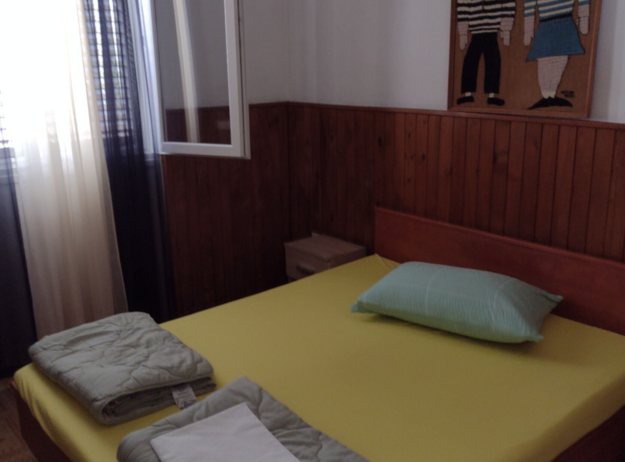 Ein kleines Zimmer mit einem Einzelbett, einem Holz-Kopfteil, einer gelben Bettdecke und einem blauen Kopfkissen. Es gibt einen kleinen Nachttisch mit einer Lampe und ein Fenster mit Vorhängen.
