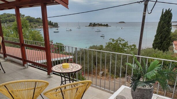 A balcony with wicker chairs and a small table, overlooking a coastal area with boats and greenery.
