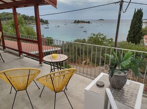Ein Balkon mit Korbstühlen und einem kleinen Tisch, mit Blick auf eine Küstenlandschaft mit Booten und viel Grün.