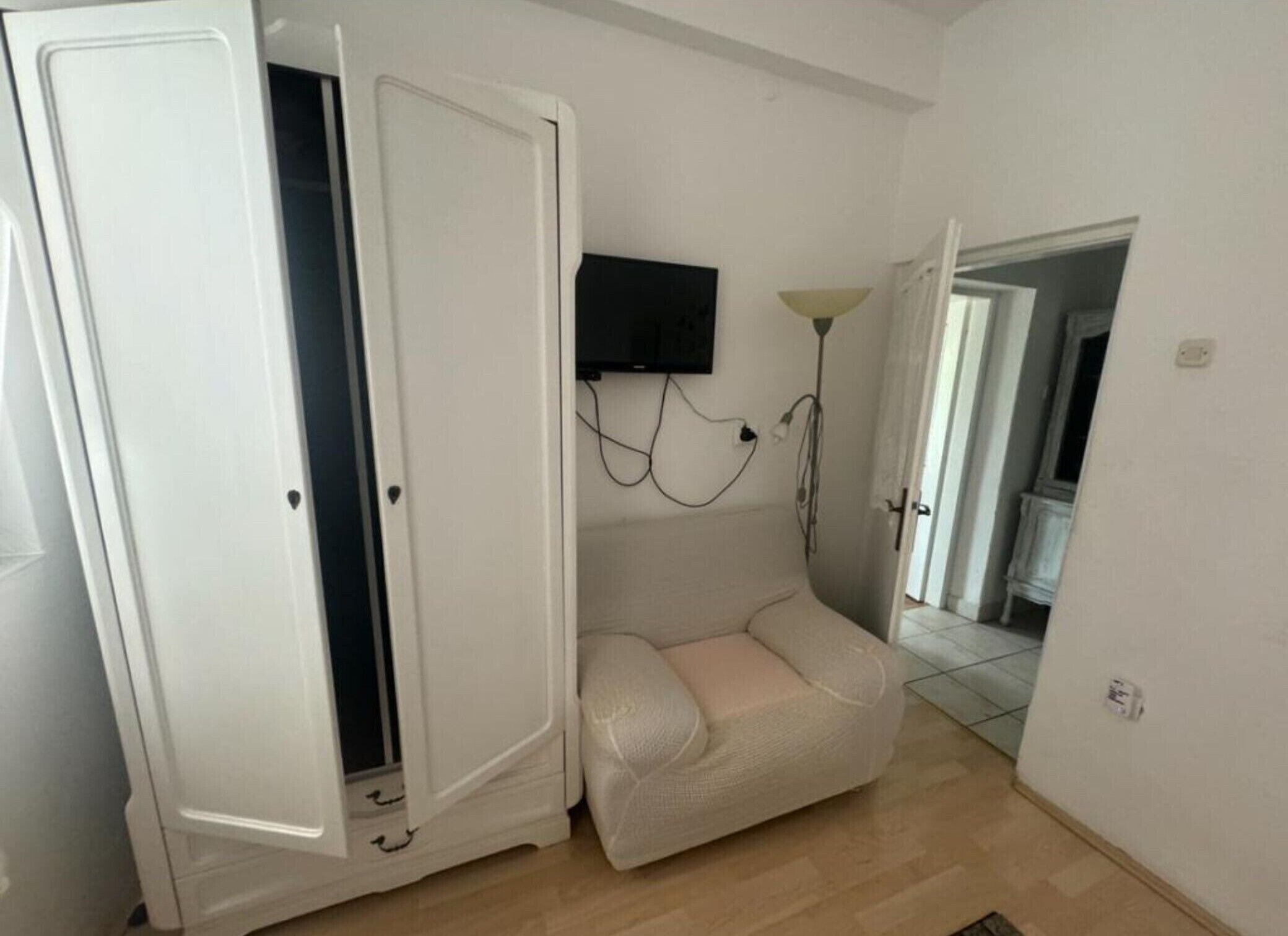 A room with a white wardrobe, a beige sofa, a mounted flat-screen TV, and a lamp.