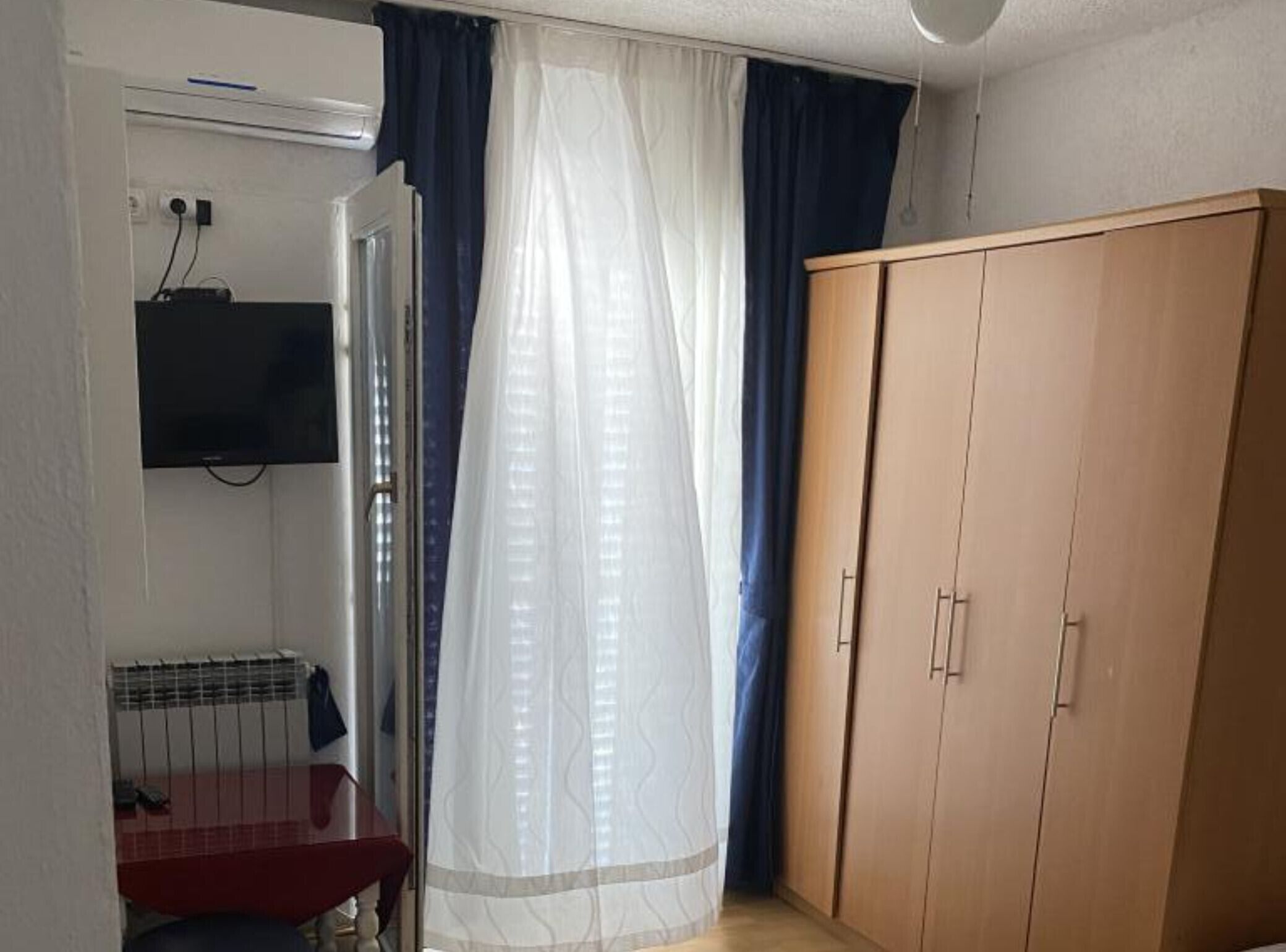 A room with a wooden wardrobe, a television mounted on the wall, and a window with sheer curtains.