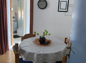 A dining area with a round table covered by a patterned tablecloth, a vase with green plants, and a clock on the wall.