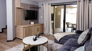 Un salon moderne doté d’un téléviseur à écran plat, d’un meuble en bois, d’une table basse ronde, d’un canapé foncé et d’une porte-fenêtre coulissante donnant sur un espace extérieur.