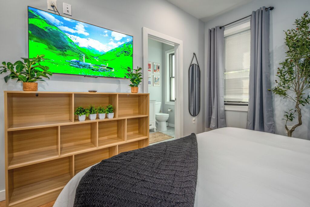 A bedroom with a bed, a TV mounted on the wall, a wooden shelf with plants, a window with curtains, and a door leading to a bathroom.