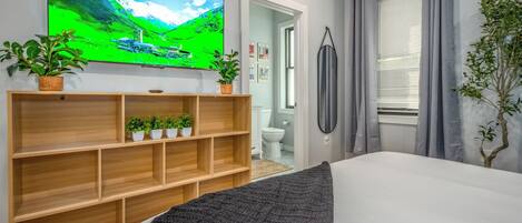 A bedroom with a bed, a TV mounted on the wall, a wooden shelf with plants, a window with curtains, and a door leading to a bathroom.