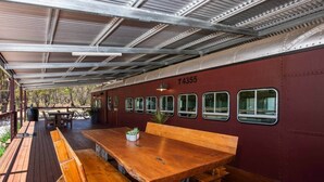 A covered outdoor dining area with a wooden table and benches, a red wall with windows, and a metal roof structure. - Red Rattler Retreat, Windeyer, Mudgee (Windeyer)