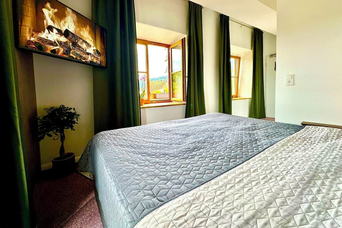 A hotel room with a large bed, a painting on the wall, a potted plant, and a window with curtains.