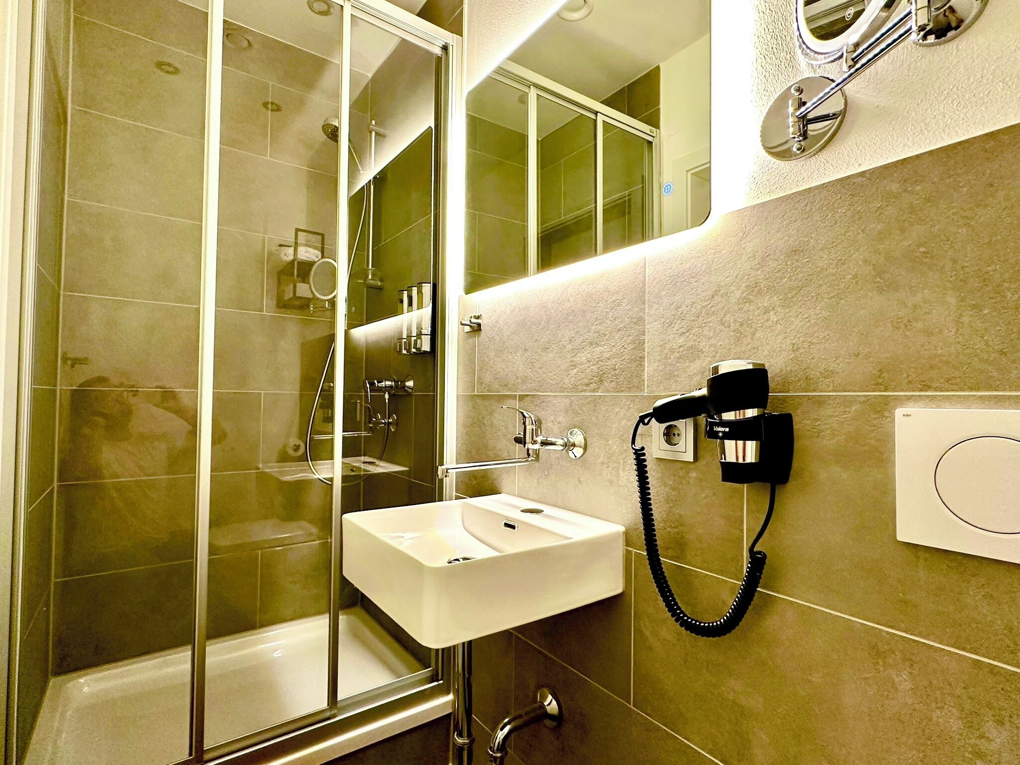 Un bagno moderno con doccia vetrata, lavandino bianco e asciugacapelli montato a parete.
