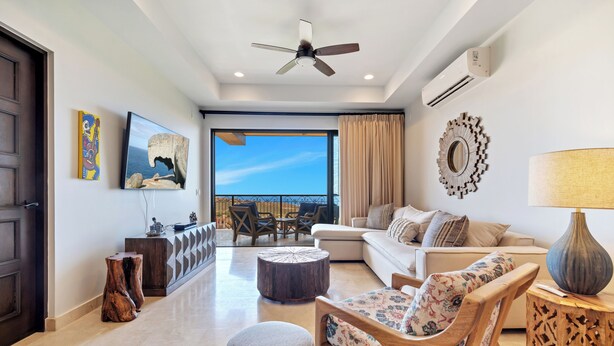 A spacious living room with a large window, a ceiling fan, and a view of the sea.
