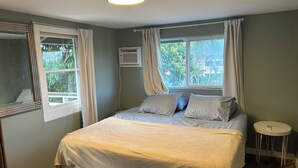 Ein Schlafzimmer mit Bett, Nachttisch, Fenster mit Vorhängen und einer Klimaanlage.