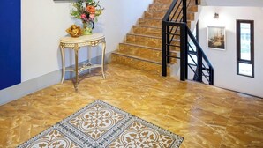 Ein geräumiger Raum mit Fliesenboden, einer Treppe mit schwarzen Geländern, einem kleinen Tisch mit Blumen in einer Vase und einem gerahmten Bild an der Wand.