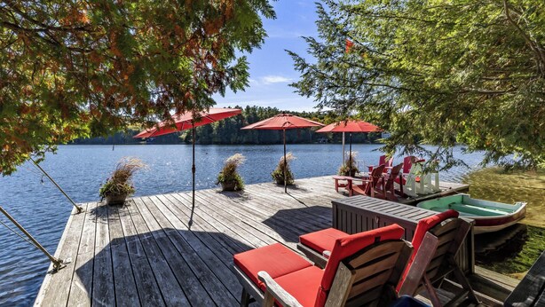 A wooden deck with red lounge chairs and umbrellas overlooking a calm lake, with boats and potted plants nearby.