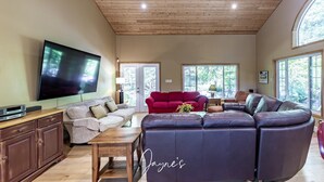 A spacious living room with a wooden ceiling, a flat-screen TV, a red sofa, a dark leather sectional, and large windows offering a view of trees.
