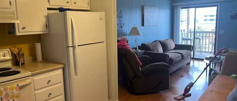 A compact kitchen with a refrigerator, stove, and cabinets. There is a dining area with a table and chairs, and a living room with a sofa and a window.