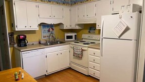 Una cocina con armarios blancos, microondas, estufa, refrigerador y una mesa de madera con algunos objetos encima.