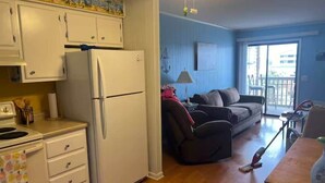 Cocina compacta con refrigerador, estufa y gabinetes blancos. Se observa un comedor con mesa y sillas, y un sofá en la sala.