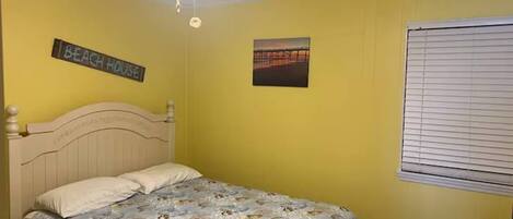 Une chambre avec un lit, un ventilateur de plafond, une fenêtre avec des stores et un panneau mural sur lequel on peut lire : « MAISON DE PLAGE ».