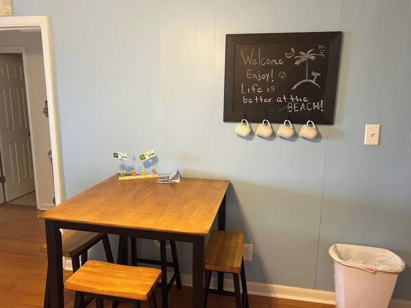 A dining area with a wooden table and benches, a chalkboard with a beach-themed message, and a trash can.