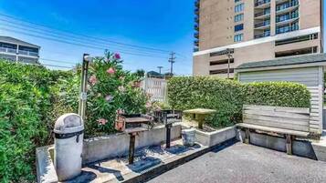 A residential area with a paved pathway, a bench, a trash bin, and a building with multiple floors.