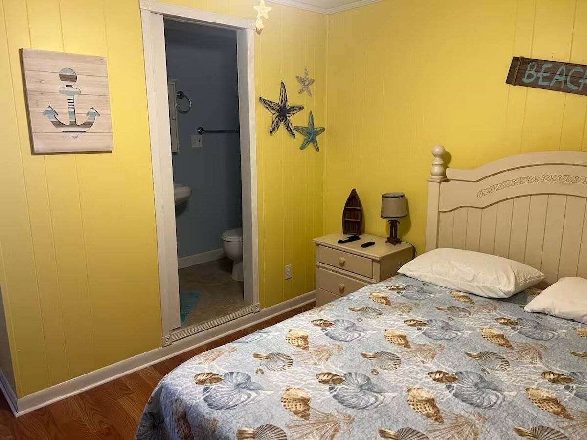 A bedroom with a bed, a nightstand, a lamp, and a door leading to a bathroom.