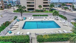 Una piscina rectangular con un bordeado y rodeada de una valla, ubicada en una zona residencial con edificios al fondo.