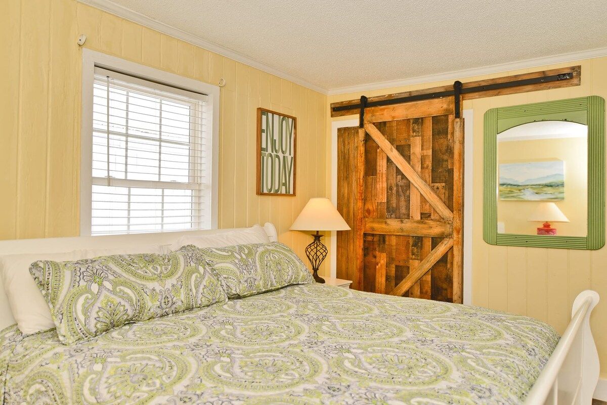 A bedroom with a bed, a window with blinds, a wooden door, a lamp, and a framed picture on the wall.