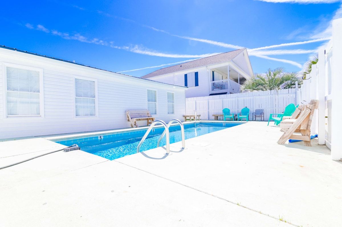 A residential swimming pool with a white house in the background, a fenced yard, and a clear sky.
