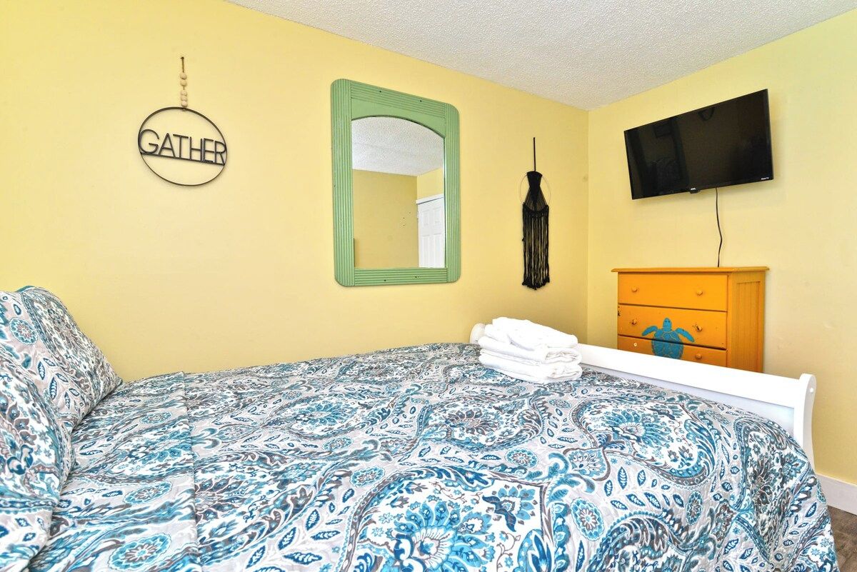 A room with a bed, a green-framed mirror, a wall-mounted TV, and a wooden dresser with drawers.