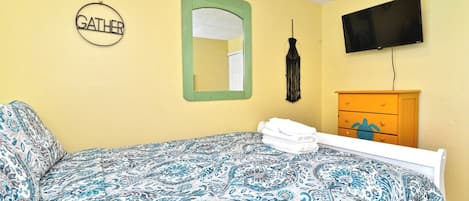 Una habitación con cama, un espejo con marco verde, televisor de montaje en pared y cómoda de madera con cajones.