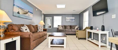 A living room with a brown sofa, a white coffee table, a grey sofa, a TV mounted on the wall, and a painting of a boat.